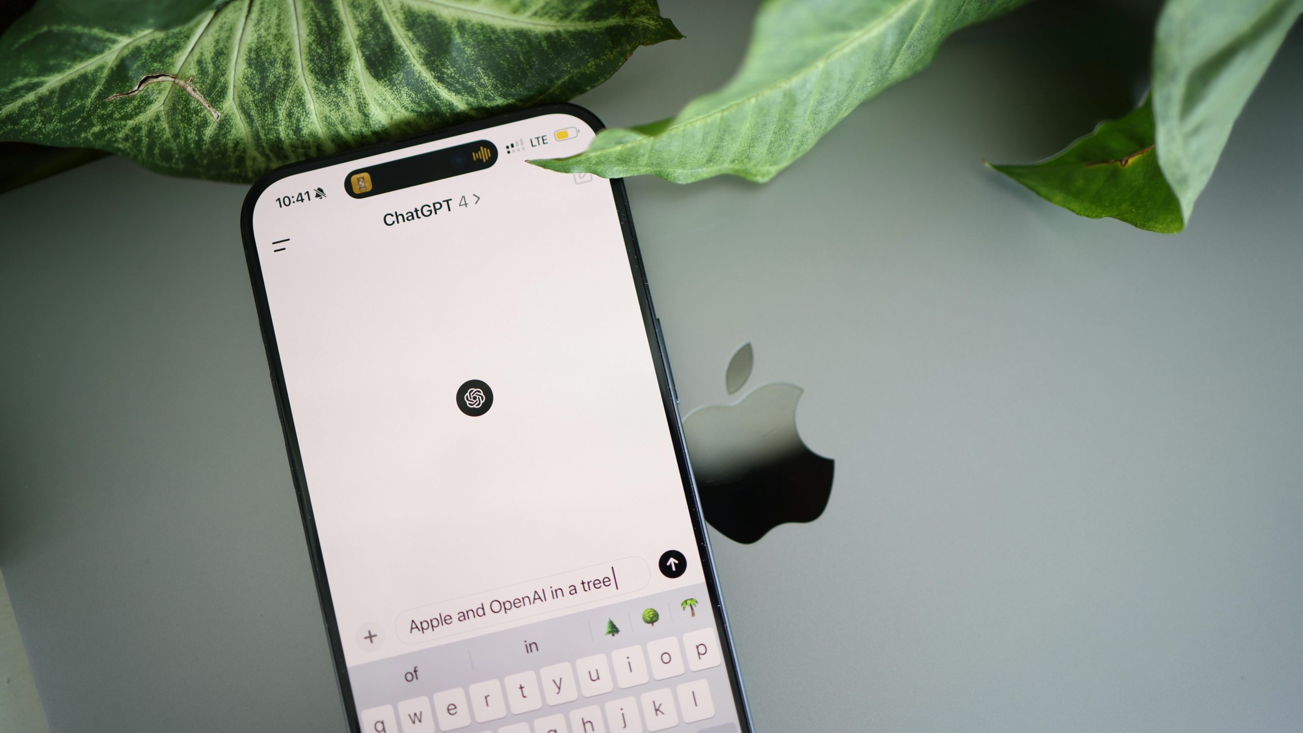 A smartphone shows a ChatGPT interface placed on an Apple laptop in a leafy environment.