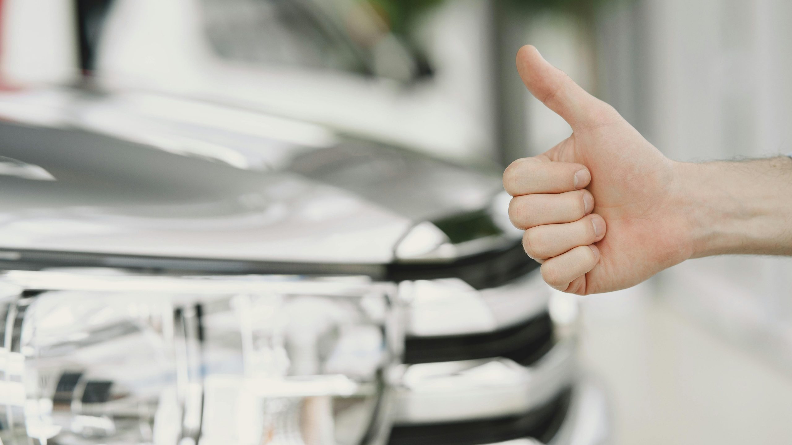 Close-up of a thumbs up gesture next to a car headlight, symbolizing approval or satisfaction.
