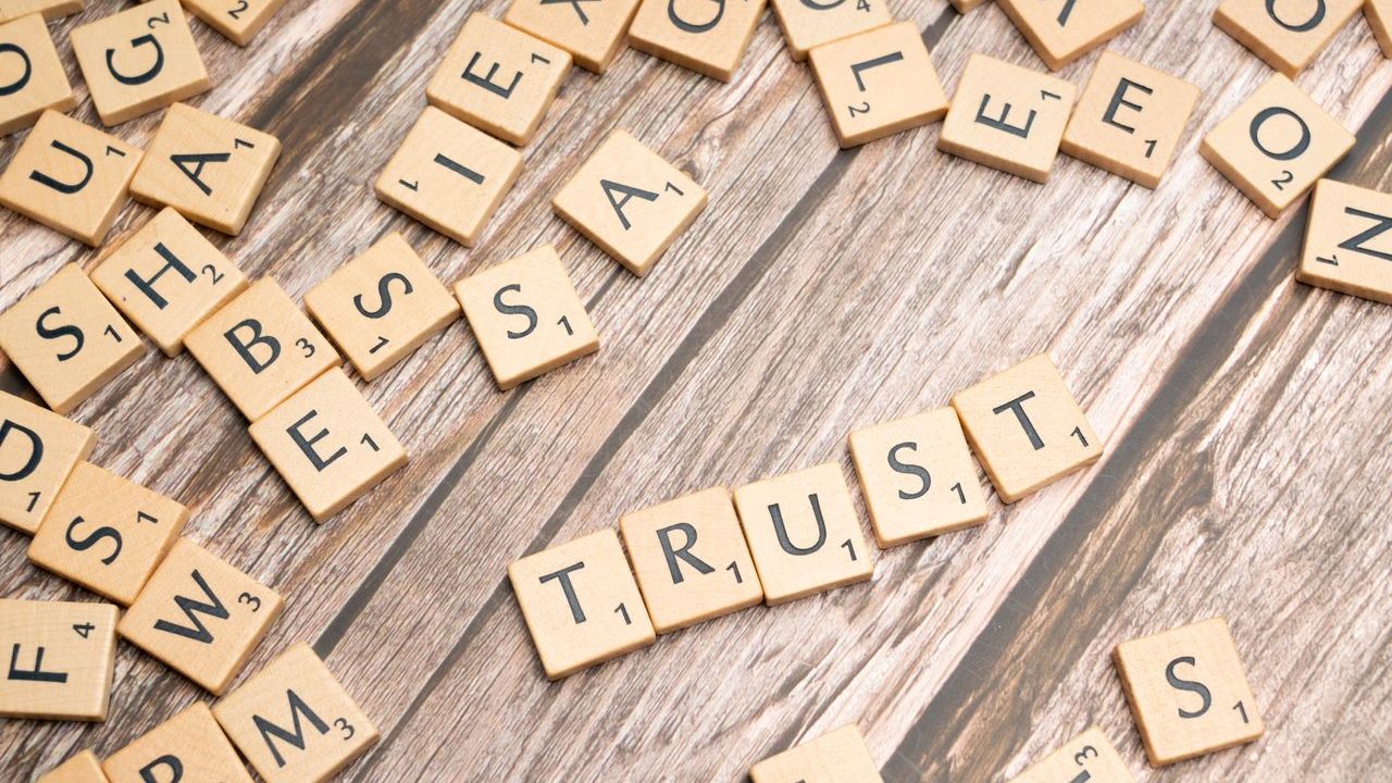 Scrabble letter tiles form the word 'Trust' on a wooden table, surrounded by scattered tiles.