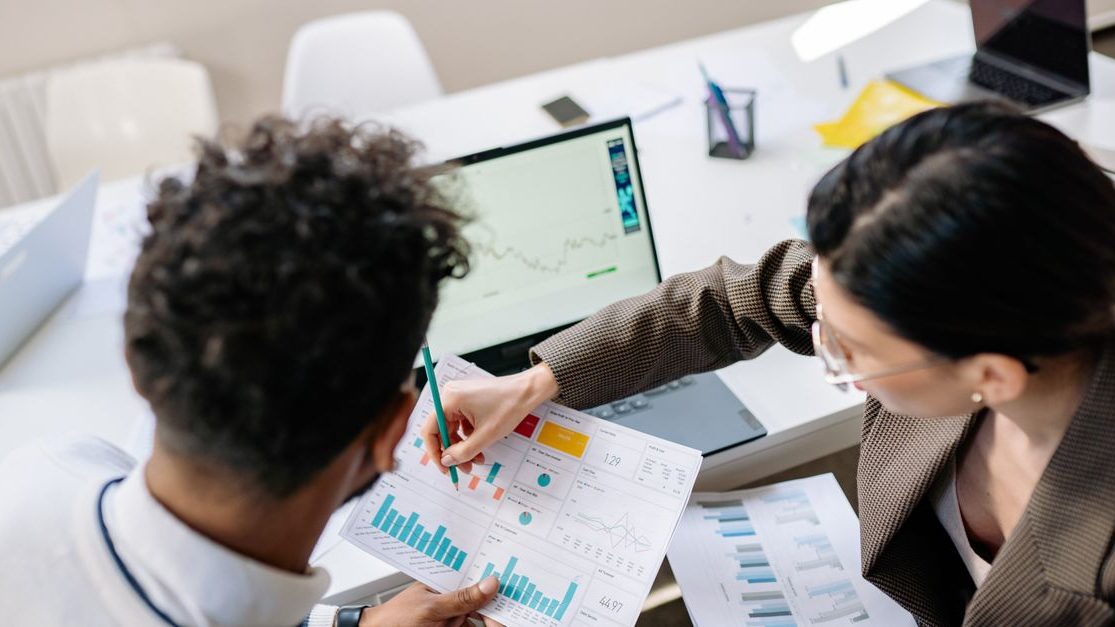 Two colleagues in a meeting room discussing financial charts and graphs on a laptop and paper.